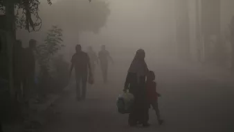 L'image montre une scène urbaine plongée dans une atmosphère brumeuse ou poussiéreuse. Dans le premier plan, une femme portant un hijab tient la main d'un jeune garçon. Ils semblent marcher ensemble le long de la rue. En arrière-plan, d'autres silhouettes de personnes sont visibles, floues par la brume, tandis que des arbres et des structures urbaines apparaissent à peine à travers la poussière. L'ambiance semble calme mais chargée d'un certain mystère à cause de la visibilité réduite.