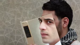 L'image présente un homme qui tient un livre devant lui, mais le livre est en quelque sorte découpé pour révéler son visage à travers la couverture. L'homme a un regard pensif et porte une écharpe à motifs. Le fond de l'image est texturé, avec un motif géométrique léger, ce qui renforce l'aspect artistique de la composition. Cela crée une impression visuelle intrigante, mêlant la lecture et l'identité.
