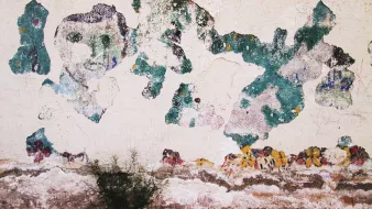 L'immagine sembra rappresentare un muro con intonaco danneggiato. La superficie è cosparsa di macchie di diversi colori, in particolare tonalità di verde e blu, che si mescolano a una base di colori più chiari e scuri. Alcuni tratti di vernice sembrano essere sbiaditi o scrostati, rivelando strati sottostanti e creando un effetto di usura nel tempo. In basso, si intravede una piccola pianta che cresce sulla superficie. L'insieme conferisce un senso di abbandono e di storia.