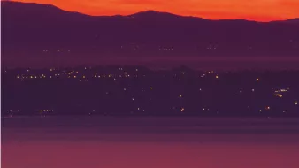 L'image présente un coucher de soleil spectaculaire, où le ciel est teinté de rouge et d'orange lumineux. En arrière-plan, on aperçoit des collines sombres, tandis qu'à l'avant-plan, un cargo navigue sur l'eau calme. Les lumières d'une ville ou d'un port scintillent le long de la côte, ajoutant une touche de vie à cette scène paisible.