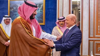 L'image montre deux hommes en costume, engagés dans une interaction amicale, probablement lors d'une rencontre officielle. L'un des hommes porte un habit traditionnel avec un châle. L'arrière-plan présente un décor élégant, typique d'un palais, avec des éléments architecturaux raffinés. D'autres personnes en tenue formelle sont visibles, suggérant un contexte diplomatique ou cérémoniel.