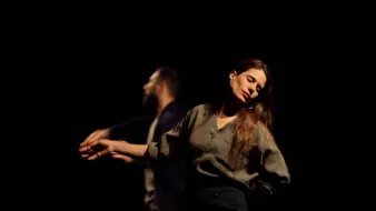Deux danseurs se déplacent sur scène dans une lumière tamisée. Une femme, les yeux fermés et une expression de sérénité, se tient légèrement en avant, tandis qu'un homme, derrière elle, semble effectuer un mouvement fluide avec ses bras. L'atmosphère est intimiste et expressive.