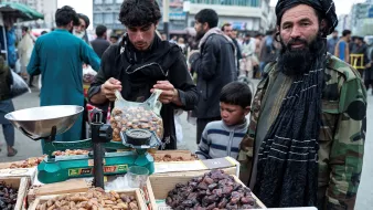 La imagen muestra un mercado al aire libre en un área concurrida. En el primer plano, hay un hombre de barba con un turbante que está de pie junto a una balanza y cajas llenas de dátiles. Otro hombre, que parece estar pesando los dátiles, está en el proceso de seleccionar los productos. También hay un niño pequeño cerca del hombre con la barba. El ambiente es activo, con varias personas en el fondo, lo que sugiere un bullicio típico de un mercado local. Las cajas de productos destacan por su variedad y colorido.