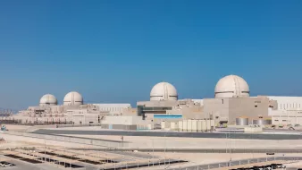 L'image montre une centrale nucléaire moderne avec plusieurs grands dômes blancs sur le toit. La structure est entourée d'un paysage clair, avec un ciel bleu sans nuages. À l'avant-plan, on peut voir des bâtiments de différentes tailles, ainsi qu'une zone de stationnement. L'ensemble de la scène dégage une impression de technologie avancée et de robustesse.