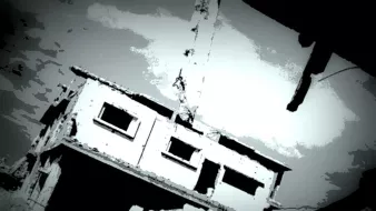 L'image montre un bâtiment en ruine, partiellement détruit, avec des murs endommagés et une atmosphère sombre. La structure semble en déséquilibre, suspendue ou penchée, ce qui renforce l'impression de dévastation. Le ciel est nuageux, ajoutant une tonalité dramatique à la scène. Les teintes sont principalement en noir et blanc, créant un effet visuel frappant.