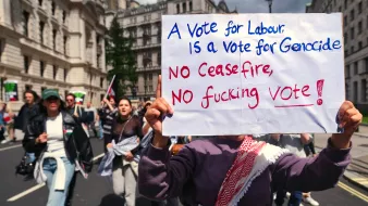 L'image montre une manifestation où des personnes défilent dans la rue, tenant des pancartes. Au premier plan, une personne tient une grande affiche avec un message fort. La pancarte indique que voter pour un certain parti est associé à des accusations graves, et elle appelle à ne pas voter sans un cessez-le-feu. Les manifestants semblent passionnés et engagés, exprimant leur désaccord par des slogans. L'ambiance est empreinte de tension et d'émotion.