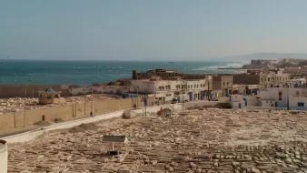 L'image montre un panorama d'une ville côtière, probablement située au bord de la mer Méditerranée. Au premier plan, on peut voir un cimetière avec de nombreuses tombes en pierre, disposées de manière régulière. À l'arrière-plan, il y a des bâtiments blancs, typiques des architectures méditerranéennes, qui bordent la côte. La mer s'étend à l'horizon, avec des vagues visibles, et il semble y avoir une légère brume sur l'eau. La scène dégage une ambiance paisible et sereine, avec une lumière douce évoquant l'heure du soir ou du matin.