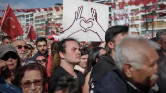 La imagen muestra una multitud reunida en un evento, posiblemente una manifestación o celebración. En primer plano, hay un hombre que sostiene un cartel en forma de corazón, dibujado con las manos. La multitud parece diversa y está vestida con ropa casual. Al fondo, hay banderas rojas con la insignia de Turquía, lo que sugiere un contexto nacional. La atmósfera parece ser de unidad y orgullo colectivo.