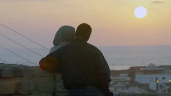 L'image montre un couple assis côte à côte, observant le coucher de soleil. L'un des membres du couple porte un hijab, tandis que l'autre est vêtu d'une veste sombre. Ils semblent se rapprocher l'un de l'autre, créant une atmosphère intime. En arrière-plan, on peut voir la mer, et le ciel est teinté de couleurs douces, avec le soleil se couchant à l'horizon. Les bâtiments sont légèrement visibles en bas, ajoutant à la scène romantique.