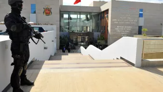 The image depicts a heavily armed police officer or security personnel standing at attention outside a building. The officer is dressed in black tactical gear and is holding a firearm. In the background, there is a modern structure with a large entrance, and the Moroccan flag is prominently displayed on top. The building appears to be a government facility, likely related to security or law enforcement, as indicated by the presence of the armed guard. The setting features stairs leading up to the entrance, with a clean, contemporary design.