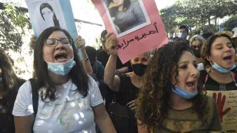 در این تصویر، گروهی از زنان و مردان در حال برگزاری یک تظاهرات هستند. آن‌ها پلاکاردهایی در دست دارند که تصاویری از افراد مختلف بر روی آن‌ها چاپ شده است. برخی از شرکت‌کنندگان به شدت فریاد می‌زنند و احساسات خود را ابراز می‌کنند. اکثر افراد ماسک به چهره دارند، که به خاطر وضعیت بهداشتی ممکن است باشد. فضای اطراف شلوغ به نظر می‌رسد و به وضوح نشان‌دهنده یک حرکت اجتماعی است.