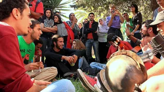 L'image montre un groupe de personnes rassemblées dans un environnement extérieur, probablement dans un parc ou un jardin. Des jeunes, assis sur l'herbe, semblent participer à une activité musicale, avec des instruments comme une guitare et des percussions. Certains d'entre eux sont engagés dans une discussion animée. En arrière-plan, d'autres personnes regardent ou écoutent, créant une atmosphère conviviale et collective. Les expressions des participants varient, allant de la concentration à l'amusement.