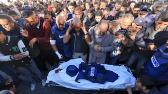 L'immagine mostra un gruppo di persone riunite in un'atmosfera di grande dolore e rispetto. Al centro, un corpo avvolto in un lenzuolo bianco è accompagnato da un giubbotto con la scritta "PRESS", suggerendo che si tratta di un giornalista. I presenti esprimono il loro cordoglio, alcuni con le mani giunte e altri con espressioni di tristezza. Il contesto sembra essere un evento commemorativo o funerario, evidenziando la gravità della situazione e il rispetto verso la persona deceduta.