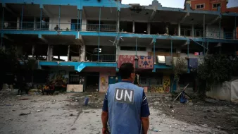The image depicts a scene of destruction, likely from a conflict area. In the foreground, a person wearing a blue vest with "UN" printed on it stands, facing a damaged building with shattered windows and debris scattered around. The building has signs of neglect, with graffiti and remnants of what may have been a children's play area. People can be seen in the background, possibly assessing the situation or providing assistance. The overall atmosphere conveys a sense of urgency and the aftermath of turmoil.