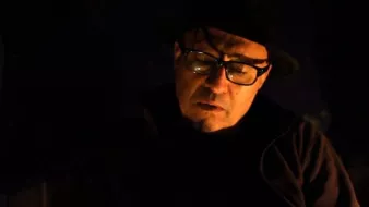 L'image montre un homme avec des lunettes et un chapeau. Il semble pensif et est plongé dans une lumière tamisée, ce qui crée une atmosphère sombre et introspective. Son expression faciale suggère qu'il est en train de réfléchir profondément ou de vivre un moment d'émotion.
