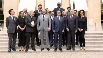 L'image montre un groupe de personnes posant sur des marches devant un bâtiment, probablement lors d'un événement officiel. Au centre, deux hommes se tiennent côte à côte, avec des costumes élégants. Les autres membres du groupe sont habillés de manière formelle ou semi-formelle et sourient pour la photo. L'ambiance semble conviviale, et l'un des hommes à l'arrière fait un signe de paix avec sa main. Les lieux sont lumineux et semblent bien entretenus, ajoutant à l'atmosphère de l'événement.