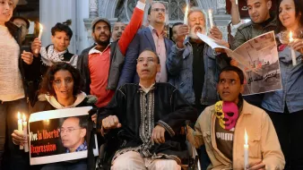 L'immagine mostra un gruppo di persone radunate in un ambiente urbano, apparentemente in un momento di protesta o di vigilanza. Alcuni partecipanti tengono in mano candele accese, mentre altri mostrano cartelli e manifesti con slogan che richiamano il tema della libertà di espressione. Tra le persone c'è un uomo in sedia a rotelle, che sembra essere al centro dell'attenzione. L'atmosfera è di solidarietà e impegno per i diritti civili.