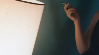L'image montre un intérieur avec une lumière douce émise par une lampe. Une personne, située à gauche, tient un objet à la main qui semble être une cigarette. La décoration murale est d'une teinte douce, créant une atmosphère intime et relaxante. La lumière de la lampe joue sur les surfaces, ajoutant une ambiance chaleureuse à la scène.