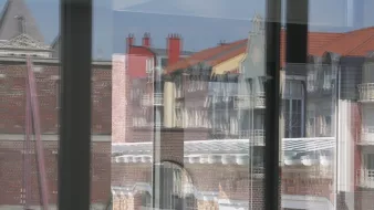 این تصویر نمایی از ساختمان‌ها را از طریق شیشه‌های یک ساختمان دیگر نشان می‌دهد. شیشه‌ها باعث ایجاد انعکاس‌هایی از بیرون می‌شوند که موجب می‌شود جزئیات معماری و رنگ‌های دیوارها و بام‌ها به خوبی نمایان شود. در پس‌زمینه، سقف‌های شیب‌دار و ساختمان‌های مختلف با طراحی‌های متفاوت دیده می‌شود، و نور آفتاب به تصویر جلوه‌ای جذاب می‌بخشد. فضای کلی تصویر حالت مدرنی دارد در حالی که عناصر تاریخی نیز در آن قابل مشاهده است.