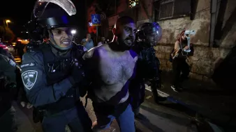L'image montre des policiers en uniforme, portant des casques et des protections, en train de maîtriser un homme. L'homme est torse nu et semble être en train d'être arrêté. L'environnement nocturne, avec des lumières et des bâtiments en arrière-plan, suggère une situation de tension, potentiellement liée à des manifestations ou des troubles civils. Les expressions et la posture des policiers indiquent une approche ferme envers la situation.