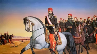 L'image représente un groupe de cavaliers en tenue traditionnelle, menés par un homme à cheval en avant-plan. Cet homme, qui semble être une figure d'autorité, est vêtu d'un uniforme orné et porte un chapeau distinctif. Il est monté sur un cheval gris, tandis qu'un groupe d'hommes sur des chevaux bruns et noirs le suit derrière lui. Le paysage est désertique, avec des montagnes en arrière-plan et un ciel aux teintes chaudes, suggérant le lever ou le coucher du soleil. Cette scène dégage une impression de mouvement et de puissance.