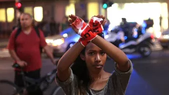 L'image montre une jeune femme qui se tient debout au milieu d'une rue, levant les bras avec des bandes de tissu rouge dans les mains, formant un symbole d'opposition ou de protestation. Elle semble déterminée et engagée, tandis que des véhicules et des lumières de police sont visibles en arrière-plan. Au fond, on peut apercevoir d'autres personnes, ajoutant un contexte urbain à la scène. L'atmosphère dégage un sentiment de mobilisation.