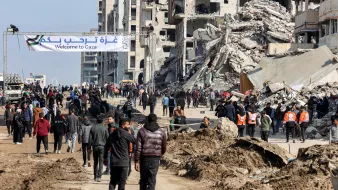 L'immagine mostra una scena in una zona colpita da conflitti, con edifici distrutti e rovine visibili sullo sfondo. Una grande insegna scritta in arabo e inglese dice "Benvenuti a Gaza". Diverse persone camminano lungo una strada, alcune indossano giubbotti arancioni, suggerendo che potrebbero essere soccorritori o volontari. L'atmosfera sembra essere quella di una comunità che cerca di ricostruirsi dopo una crisi.