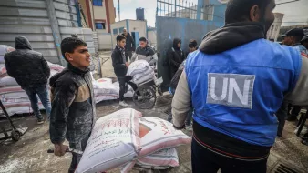 L'immagine mostra una scena di aiuto umanitario. Alcuni ragazzi e adulti sono impegnati nel trasporto di sacchi, probabilmente di cibo o materiali di prima necessità, all'interno di una zona urbana. Un ragazzo sta spingendo un carrello con sacchi, mentre altre persone, alcuni dei quali indossano giubbotti con il logo delle Nazioni Unite, sono visibili in background e stanno trasportando ulteriori carichi. La scena comunica un senso di collaborazione e supporto nella distribuzione di aiuti umanitari.