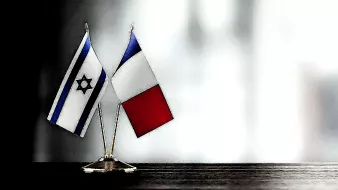 The image features two small flags positioned on a surface. One flag is the national flag of Israel, characterized by its blue and white colors and a Star of David in the center. The other flag is the national flag of France, which consists of three vertical stripes: blue, white, and red. The flags are mounted on small stands, standing together side by side, against a softly blurred background.