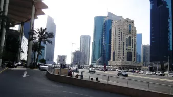 تظهر الصورة مشهداً حضرياً لمدينة حديثة، حيث تتداخل المباني الشاهقة مع تشكيلة متنوعة من التصاميم المعمارية. في الجهة اليسرى، توجد أشجار النخيل التي تضيف لمسة طبيعية على المشهد. يتواجد على الطريق مجموعة من السيارات، مما يشير إلى حركة المرور في المنطقة. السماء تظهر بلون فاتح، مما يوحي بأنها يوم مشمس. العمارات تتنوع في ارتفاعاتها وأشكالها، مما يعكس الطابع العصري للمدينة.