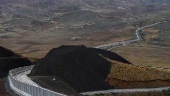 این تصویر نمایی از یک جاده در میان مناظر طبیعی خشک و کوهستانی را نشان می‌دهد. جاده به صورت منحنی و پیچ‌درپیچ از روی زمین‌های ناهموار عبور می‌کند و در سمت راست تصویر، دیواری سفیدرنگ وجود دارد که به طور راستا در حال پیشروی است. در پس‌زمینه، تپه‌ها و دامنه‌های کوهستانی با رنگ‌های ملایم و خاصی دیده می‌شوند که نمایی طبیعی و بکر را به تصویر می‌بخشند. فضای کلی تصویر به نوعی حس دورافتادگی و خلوتی را منتقل می‌کند.