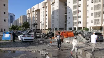 در این تصویر، یک منطقه شهری به نمایش گذاشته شده که به نظر می‌رسد به خاطر یک حادثه انفجاری آسیب دیده است. ساختمان‌ها در دو طرف خیابان قرار دارند و در میان آنها، خودروهای beschädigt و ویران‌شده به چشم می‌خورند. زباله‌ها و قطعات شکسته در سطح زمین پخش شده‌اند و نشانه‌هایی از ویرانی دیده می‌شود. چند نفر در حال قدم زدن در این مکان آسیب‌دیده هستند و به نظر می‌رسد در حال ارزیابی وضعیت هستند. همچنین، آب‌ای که ممکن است بر اثر باران یا دیگر عوامل جمع شده باشد، در بعضی نقاط خیابان دیده می‌شود. آسمان آبی و روشن به نظر می‌رسد.