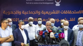 Sur l'image, un groupe d'hommes se tient devant une bannière affichant le nom d'un parti politique, le Parti de la Justice et du Développement. Les hommes portent des masques de protection, ce qui suggère un contexte de santé publique. Ils semblent s'apprêter à faire une déclaration ou une conférence de presse, entourés de microphones et de caméras. L'atmosphère semble sérieuse et professionnelle.