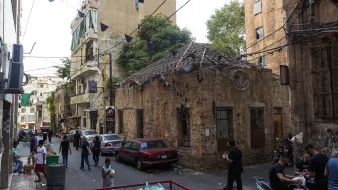 The image depicts a narrow street lined with older buildings, showcasing a blend of urban decay and lively activity. There's a mixture of people, some walking on the street and others gathered in small groups. The buildings have seen better days, with peeling paint and signs of wear. A small, weathered structure with a distinctive roof stands out against the backdrop of larger, more modern buildings. Utility wires crisscross overhead, and there's a hint of greenery from nearby trees. The scene reflects a vibrant yet gritty neighborhood atmosphere.