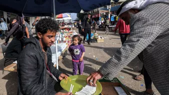 L'immagine mostra una scena di mercato all'aperto, probabilmente in una città. Un giovane uomo è seduto su un contenitore di legno, di fronte a un tavolo, mentre tiene in mano un piatto. Sta vendendo o mostrando dei prodotti, probabilmente cibo o articoli vari. Accanto a lui, un uomo anziano, vestito con indumenti tradizionali, si avvicina per prendere qualcosa dal piatto. Sullo sfondo, ci sono altre persone che passeggiano e alcune bancarelle con vari articoli in esposizione. Una bambina, vestita con una maglietta, osserva curiosa la scena. La luce del giorno illumina vividamente l'ambiente, creando un'atmosfera vivace e animata.