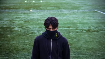 L'image montre un jeune homme se tenant sur un terrain de sport couvert d'herbe. Il porte un sweat à capuche noir et un masque noir qui couvre une partie de son visage. Son regard est intense et fixé vers l'objectif, créant une atmosphère mystérieuse. Le terrain derrière lui est bien entretenu, avec des marques blanches au sol.
