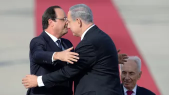 L'image montre deux hommes qui se serrent dans les bras dans un geste amical, tandis qu'un troisième homme est assis derrière eux, regardant la scène. Les deux hommes à l'avant portent des costumes, et on peut notamment voir l'un d'eux pencher la tête en signe de proximité. L'arrière-plan est basé sur une surface rouge, suggérant un événement officiel ou cérémoniel. L'atmosphère semble conviviale et respectueuse.