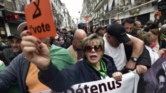 L'image montre une manifestation dans une rue d'Alger. Au premier plan, une femme tient une pancarte orange avec le mot "l'vote". Elle est entourée par d'autres manifestants, dont certains portent des drapeaux algériens. L'atmosphère semble engagée et déterminée, avec des expressions de solidarité parmi les participants. Des banderoles sont visibles, indiquant des revendications liées à la situation politique. L'environnement urbain et les bâtiments en arrière-plan témoignent d'une ambiance dynamique de protestation.