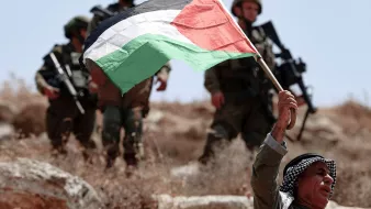 L'image montre un homme brandissant fièrement un drapeau palestinien. Il semble être en première ligne, tandis que des soldats en arrière-plan, probablement des soldats israéliens, sont visibles. Le paysage est sec et rocailleux, suggérant un environnement rural ou semi-aride. L'atmosphère semble tendue en raison de la présence militaire et du geste de protestation de l'homme.