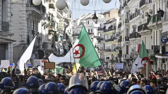 تظهر الصورة تجمعًا حاشدًا في شارع بالجزائر، حيث يحمل المتظاهرون الأعلام الجزائرية ويعبرون عن مطالبهم. يوجد في مقدمة الصورة رجال شرطة يرتدون خوذات واقية، وكأنهم يراقبون الوضع. يُلاحظ وجود لافتات ومحتجين يعبرون عن آراءهم، مما يدل على حماس واهتمام المواطنين بالقضايا المحلية. الأجواء تبدو مليئة بالحيوية والتوتر في آن واحد.