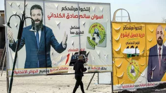 تظهر الصورة شخصًا يسير بجانب لوحات إعلانات كبيرة تتضمن صورًا وعناوين تتعلق بانتخابات. إحدى اللوحات تحتوي على صورة لرجل يُدعى سالم صادق الكلداني، والأخرى تتعلق بشخص يُدعى جليل بشير. النص في الإعلانات يتضمن عبارات تدعو الناخبين لاختيار المرشحين للمزيد من التفاعل والمشاركة السياسية. الخلفية تسلط الضوء على أجواء انتخابية تشهدها منطقة معينة.