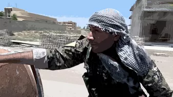 La imagen muestra a un hombre vestido con un uniforme militar y una kefía (una especie de pañuelo) en la cabeza. Él parece estar en un entorno urbano, posiblemente en un área de conflicto. El fondo incluye edificios y una zona que parece abandonada o dañada. El hombre tiene una expresión concentrada y parece estar en alerta, posiblemente observando o preparándose para algo en el entorno que lo rodea.