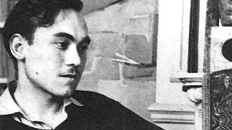 L'image présente un jeune homme assis, vêtu d'un pull noir et d'une chemise claire. Il a les cheveux foncés et porte une expression pensive, regardant légèrement à gauche. Le décor en arrière-plan suggère un intérieur avec des éléments architecturaux, bien que les détails ne soient pas très clairs. L'atmosphère de la photo semble rétro, avec un contraste marqué entre les ombres et la lumière.
