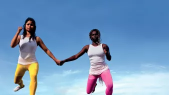 L'image montre deux femmes qui semblent flotter dans les airs, se tenant par la main. Elles portent des vêtements de sport, l'une en leggings jaunes et l'autre en leggings roses, avec des t-shirts blancs. En arrière-plan, on peut voir une ville urbaine s'étendre sous elles, avec un ciel bleu clair. Cette composition crée une sensation de liberté et de mouvement dynamique.