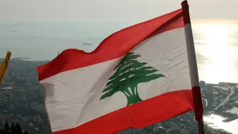 L'image montre le drapeau du Liban flottant au vent, avec un fond qui semble représenter une vue panoramique d'une ville côtière. Le drapeau se compose de trois bandes horizontales : rouge en haut et en bas, et une bande blanche au centre avec un grand cèdre vert, emblème national du Liban. L'horizon présente une mer scintillante, probablement au coucher du soleil, ce qui ajoute une ambiance paisible et naturelle à la scène.