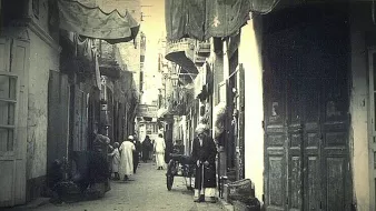 L'image montre une ruelle étroite d'une ville ancienne, probablement dans un pays arabe. Les murs des bâtiments sont ornés de tentures et de draps qui ajoutent une ambiance colorée. On peut voir des habitants, dont certains portent des vêtements traditionnels, déambulant dans la rue. Un homme semble se tenir près d'une charrette, tandis que d'autres personnes se déplacent en arrière-plan. Les portes en bois, typiques, ajoutent un charme ancien à la scène, qui évoque une atmosphère de vie quotidienne dans un vieux quartier.