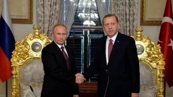 En la imagen se pueden ver a dos líderes políticos, uno de Rusia y otro de Turquía, dándose la mano en un acto formal. Ambos están vestidos con trajes oscuros y corbatas, y parecen estar en un entorno elegante, rodeados de decoraciones doradas y banderas de sus respectivos países. La atmósfera sugiere un momento de colaboración o acuerdo entre las dos naciones.
