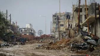 الصورة تظهر مشهدًا لمدينة مدمرة، حيث تبدو المباني متضررة بشدة، مع بقايا الهياكل المعدنية وأجزاء من الأنقاض. الطريق في المنتصف مليء بالحصى والأحجار، ويبدو غارقًا في الخراب. يظهر في الخلفية بعض الأعمدة الكهربائية التي لم تعد تعمل، مما يضيف إلى الجو القاتم والموحش للمكان. الأجواء عامة غائمة، مما يزيد من شعور الخواء واليدانة في هذا المشهد.