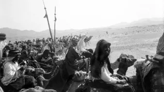 تصویری سیاه و سفید از یک کاروان در بیابان است. گروهی از افرادی که بر روی شترها سوار هستند، در حال حرکت به سمت دوربین هستند. در میان آنها زنانی با پوشش‌های سنتی و مردانی با لباس‌های مختلف دیده می‌شوند. برخی از افراد نیز پرچم‌هایی در دست دارند. پس‌زمینه تصویر شامل تپه‌های شنی و کوه‌ها است که حس فضای بیابانی را القا می‌کند. جو تصویر قدیمی و تاریخی به نظر می‌رسد.
