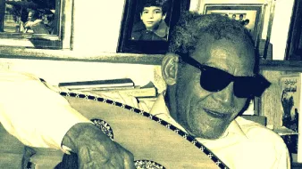 الصورة تظهر رجلًا مسنًا يرتدي نظارات شمسية ويعزف على آلة العود. يبدو سعيدًا ومسترخيًا، محاطًا بإطارات صور على الجدار خلفه. الأسلوب الذي يقدّم به العزف يعكس شغفه بالموسيقى. الأجواء تدل على لمسة من الح nostalgia، مع تفاصيل تعكس العناية والحنين إلى الماضي.